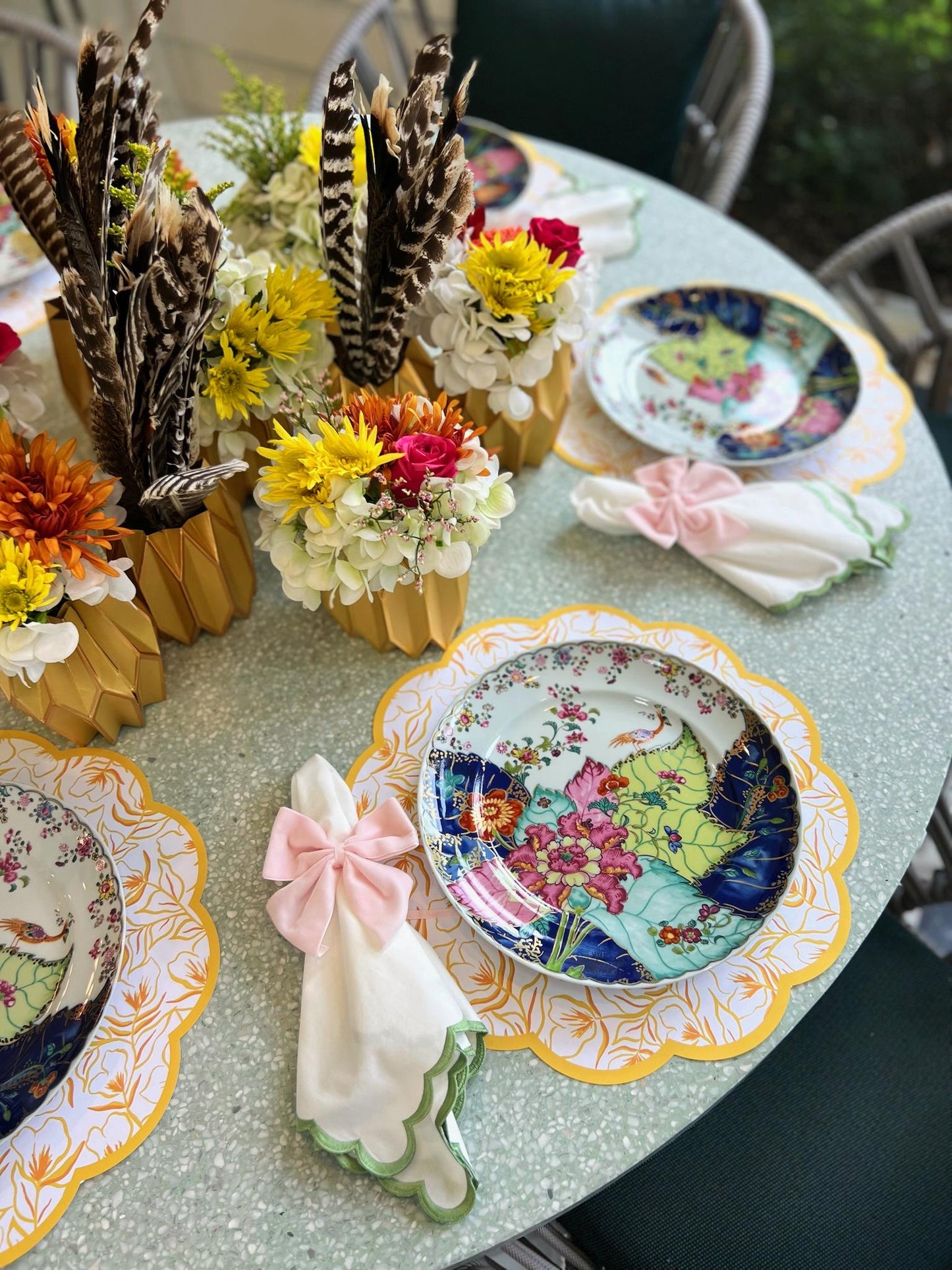 A tablescape featuring scalloped paper placemats with marigold pattern and gold border, colorful china with vibrant tobacco leaf print, and vases with faux feathers and flowers wrapped in gold paper vase sleeves