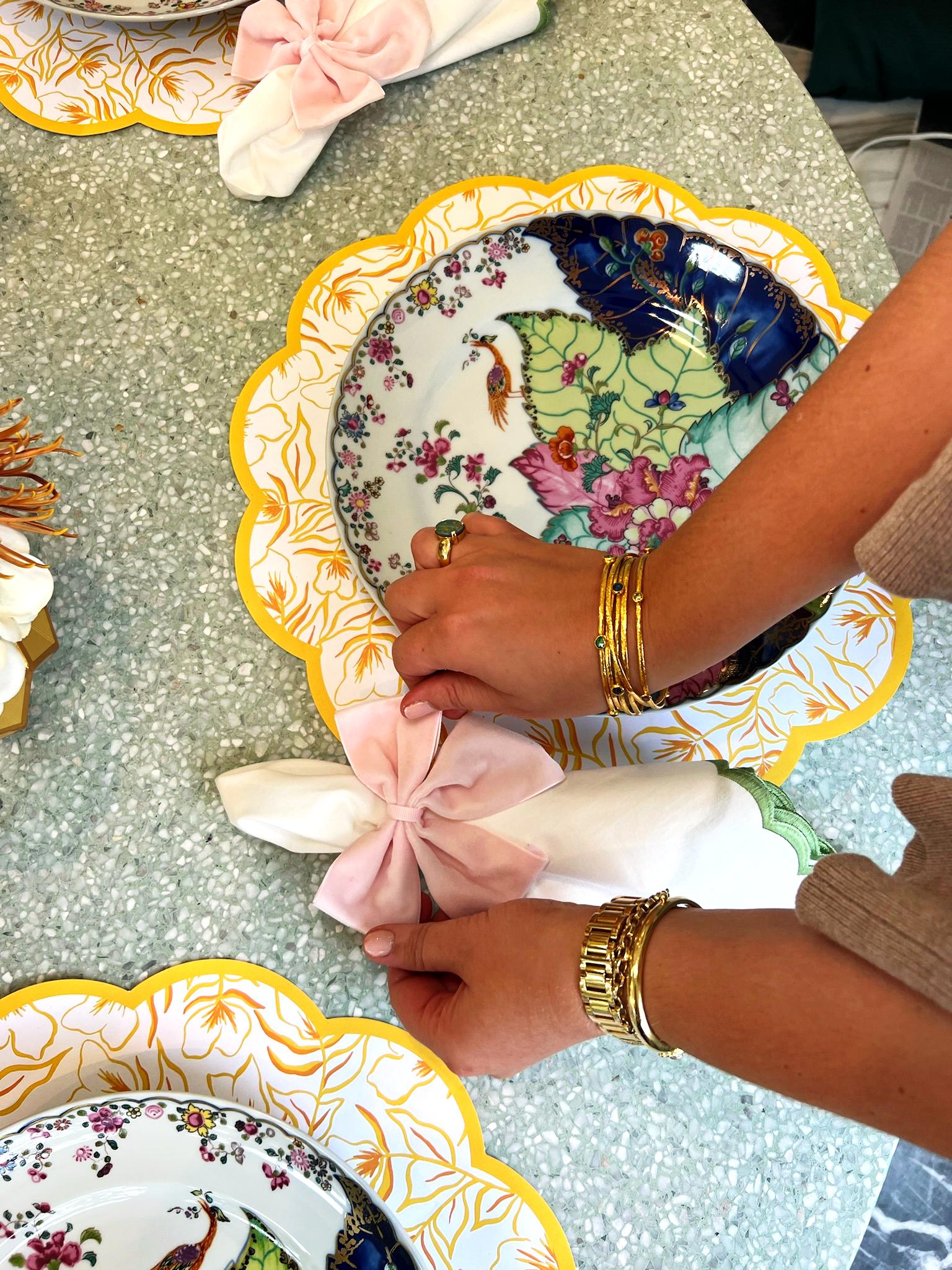 A tablescape featuring scalloped paper placemats with marigold pattern and gold border, colorful china with vibrant tobacco leaf print, and vases with faux feathers and flowers wrapped in gold paper vase sleeves