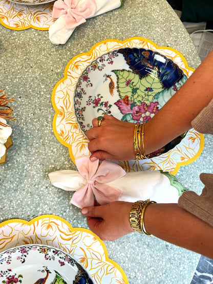 A tablescape featuring scalloped paper placemats with marigold pattern and gold border, colorful china with vibrant tobacco leaf print, and vases with faux feathers and flowers wrapped in gold paper vase sleeves