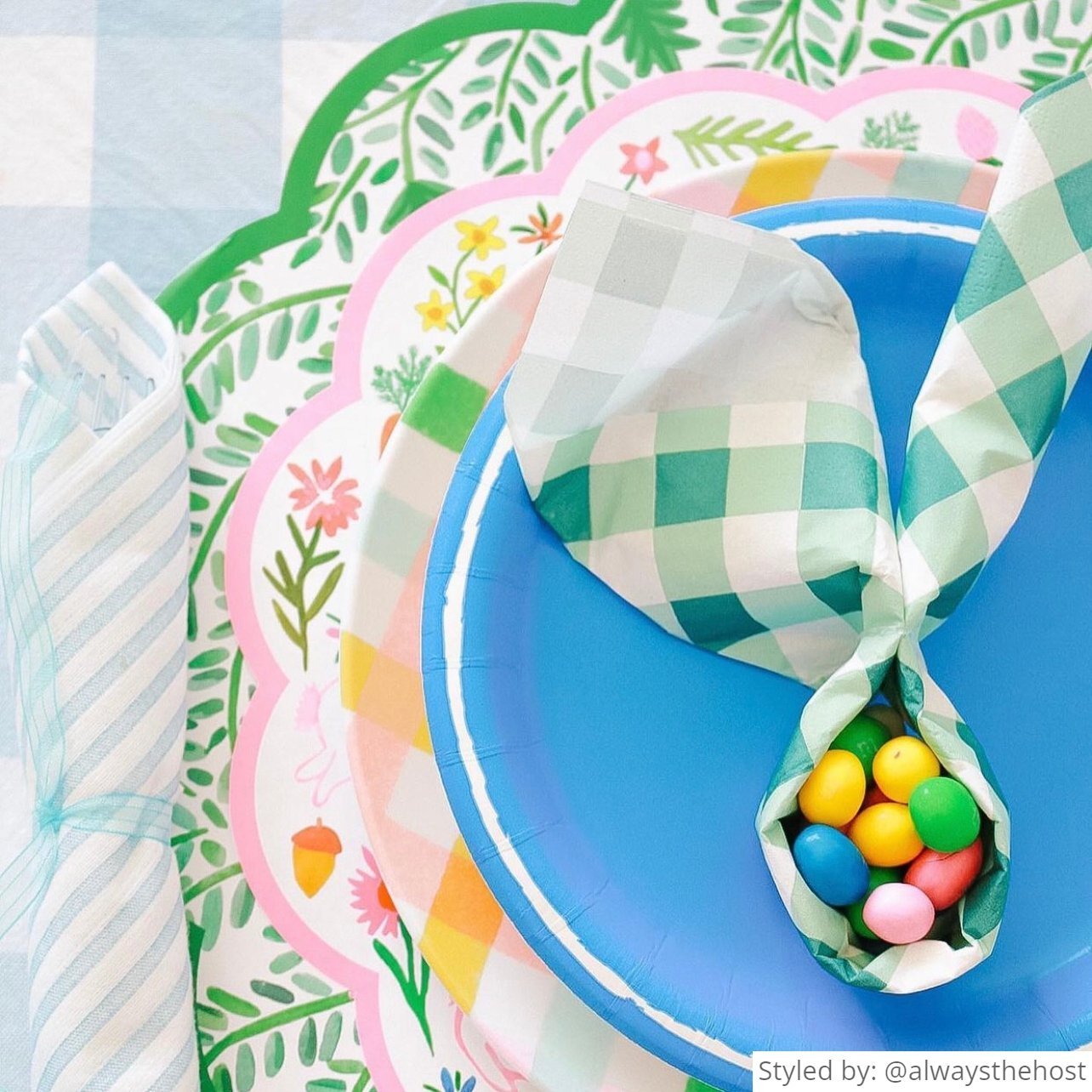 Close up of a place setting with a green gingham round scalloped placemat layered with bunny round scalloped chargers, a pastel gingham plate and a blue plate with a green gingham napkin folded into a bunny shape with colorful jelly beans