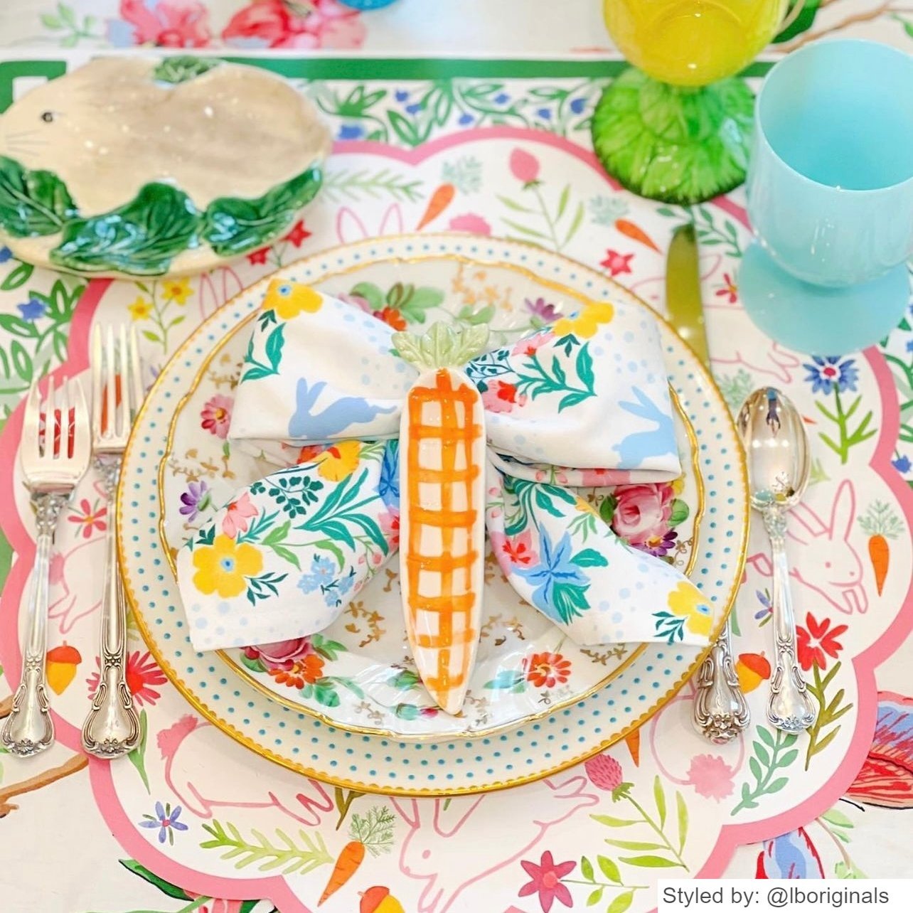 Easter place setting with a bunny scalloped round paper placemat layered with china and a floral napkin tied into a bow with a carrot shaped dish on top