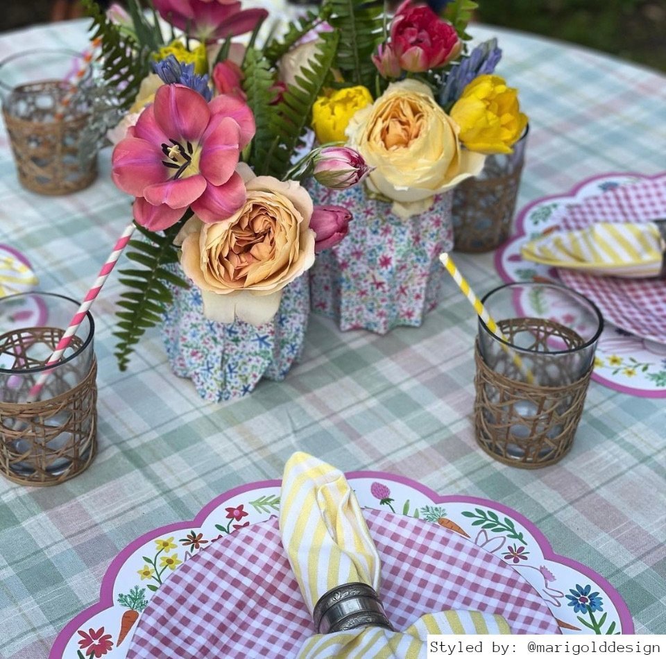 Table setting with bunny paper placemats on a plaid tablecloth with multicolored floral paper vases with spring flowers as centerpieces