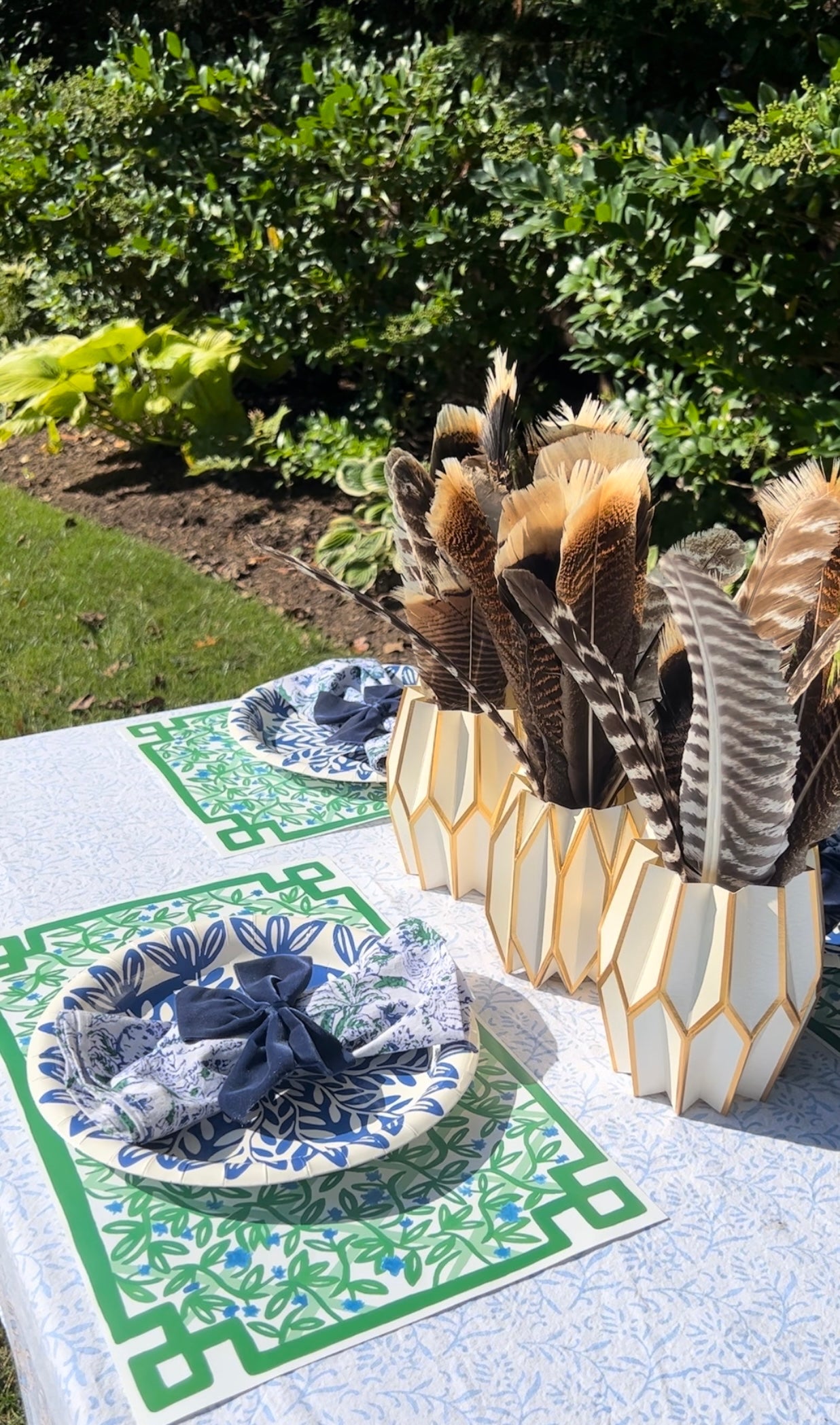 Blue and green tablescape featuring our Trellis rectangle paper placemats, blue heavy duty paper plates, blue bow napkin rings, and white and gold paper vase sleeves